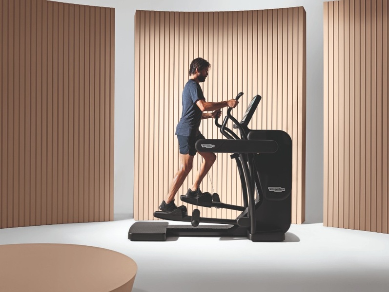 A high-quality 4:3 static image featuring a user on an Elliptical Trainer, positioned against a neutral, warm-toned ribbed backdrop.